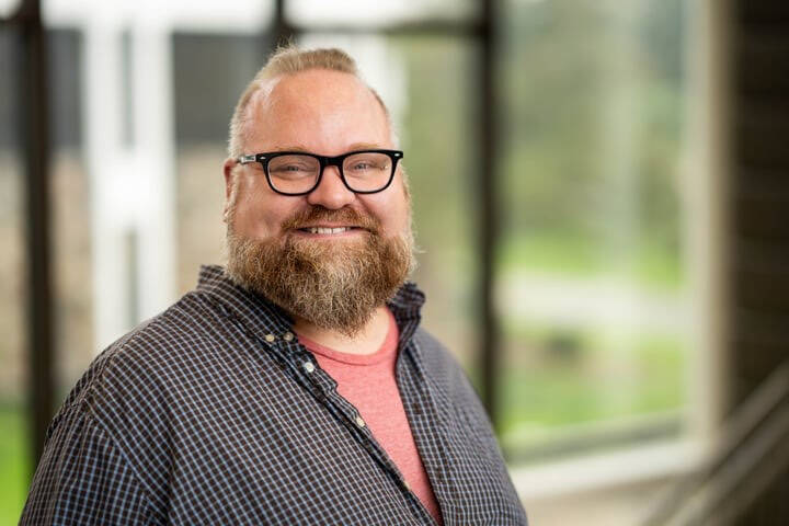 Portrait of a smiling man with a beard wearing reading glasses and a grey button down shirt, standing in front of a blurred green background.